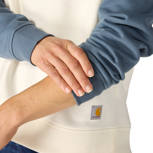 Close-up of a person wearing a Carhartt sweatshirt, showing the blue ribbed cuff of the long-sleeve layer over a cream-colored base. The Carhartt logo patch is visible on the lower front.