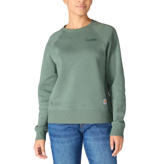 A woman wearing a sage green Carhartt crewneck sweatshirt and blue jeans, standing against a plain white background.