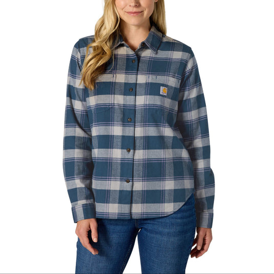A woman modeling a Carhartt long-sleeve plaid button-up shirt in blue and gray, paired with blue jeans, against a solid white background.