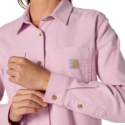 A close-up of a person wearing a light pink long-sleeve button-up shirt with a chest pocket, gold-tone buttons, and a visible Carhartt logo patch on the pocket.
