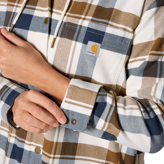 Close-up of a person wearing a Carhartt plaid hooded shirt, showing the chest pocket with the brand label, drawstrings, and the cuff being adjusted.
