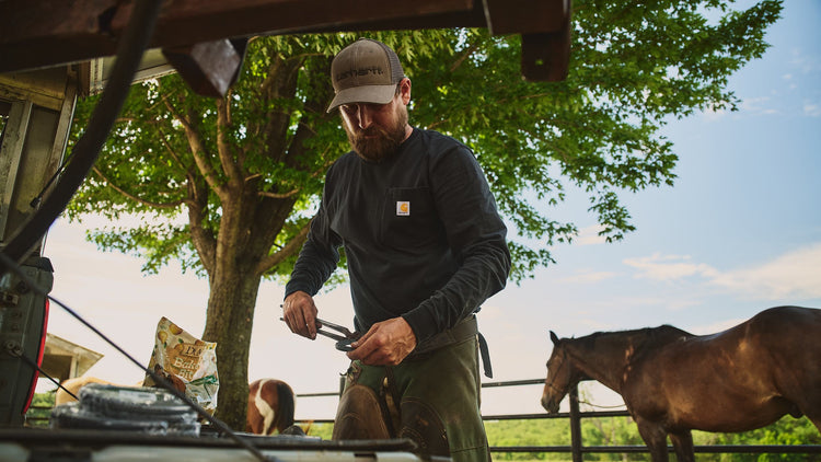 A man wearing a brown Carhartt cap and a dark long-sleeve work shirt stands outdoors in a farm setting, using a pair of hand tools near a metal container. A horse is visible in the background under a large tree.