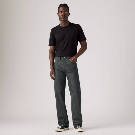 A model stands against a light gray studio background wearing a black short-sleeve crewneck t-shirt, dark gray straight-leg jeans, and black low-top sneakers with white soles.