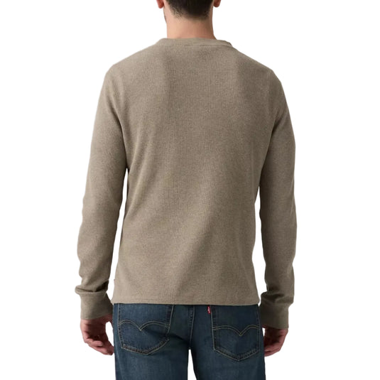 A man viewed from behind wearing a light taupe-beige long-sleeve knit sweater and dark blue jeans against a plain white studio background.