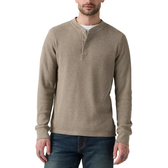A man wearing a taupe-beige long-sleeve henley sweater over a white undershirt and dark blue jeans, standing against a white studio background.