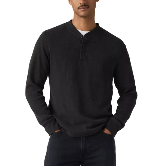 A man wearing a black long-sleeve henley shirt and dark jeans, standing against a light gray studio background.