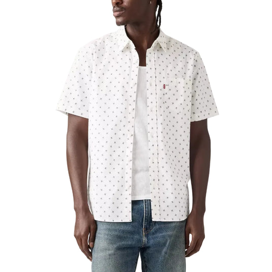 A man wearing a white short-sleeve button-up shirt with a small repeating dark pattern, layered over a white t-shirt and blue jeans, photographed against a light gray studio background.