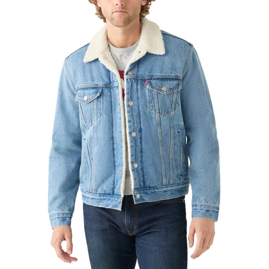 A man wearing a light blue denim trucker jacket with a cream sherpa collar, layered over a white t-shirt and dark blue jeans, against a plain white background.