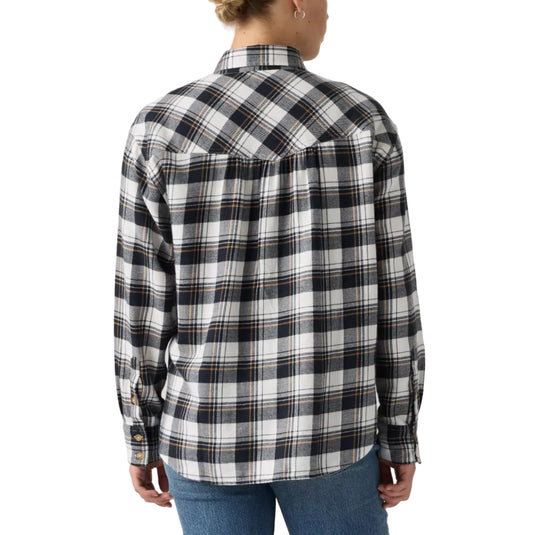 A woman viewed from the back wearing a black, white, and gray plaid flannel shirt with blue jeans, set against a plain light gray studio background.