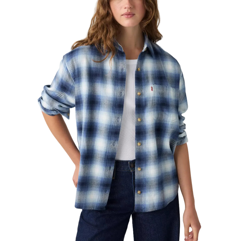 Load image into Gallery viewer, A woman modeling a blue and white plaid button-up shirt with rolled-up sleeves, worn over a white top and dark jeans, against a light gray studio background.
