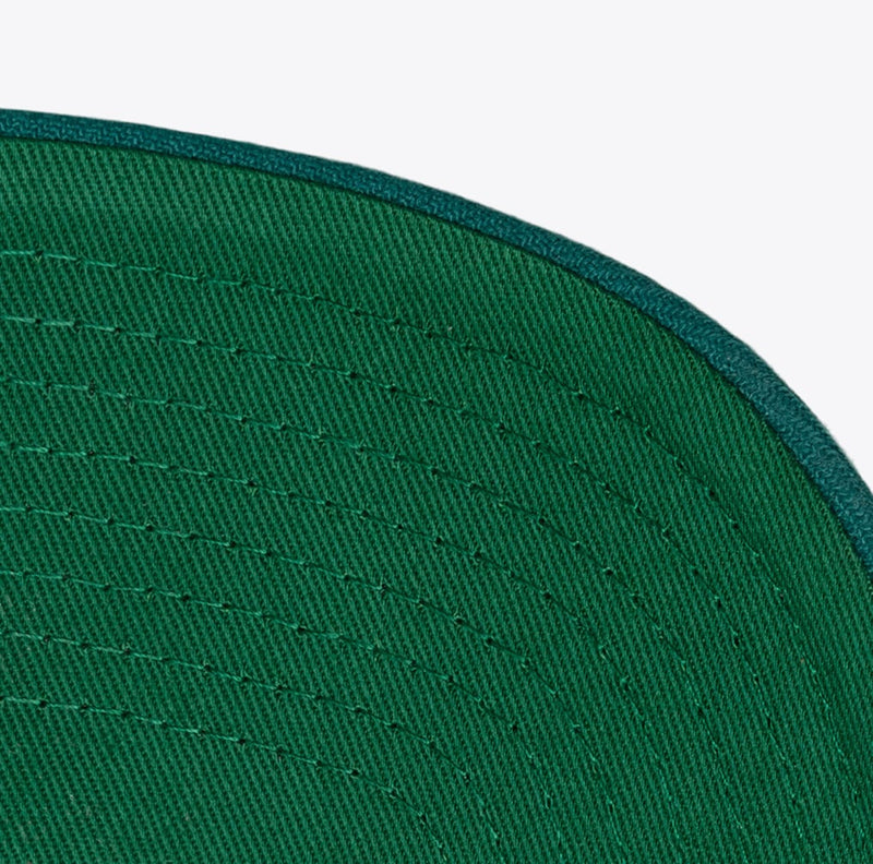 Load image into Gallery viewer, Close-up detail of a green baseball cap showing the curved brim edge, stitched fabric texture, and dark green piping against a light gray background.
