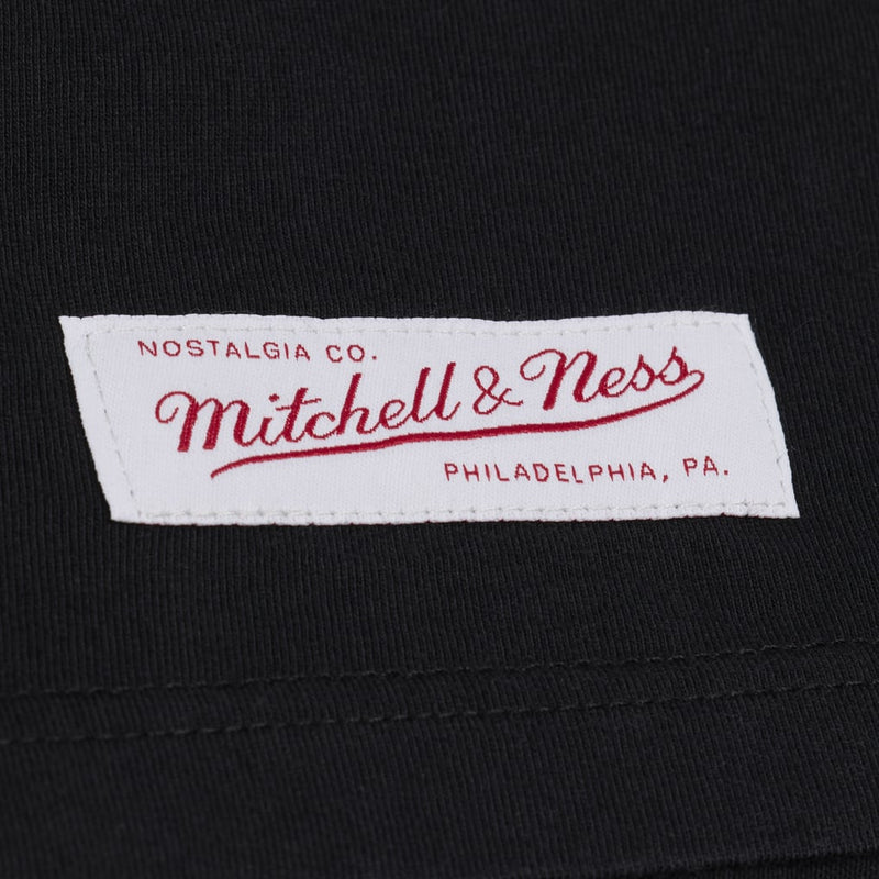 Load image into Gallery viewer, Close-up of a black knit garment with a white Mitchell &amp; Ness brand label sewn onto the chest. The label features red embroidered text reading 'Nostalgia Co.

