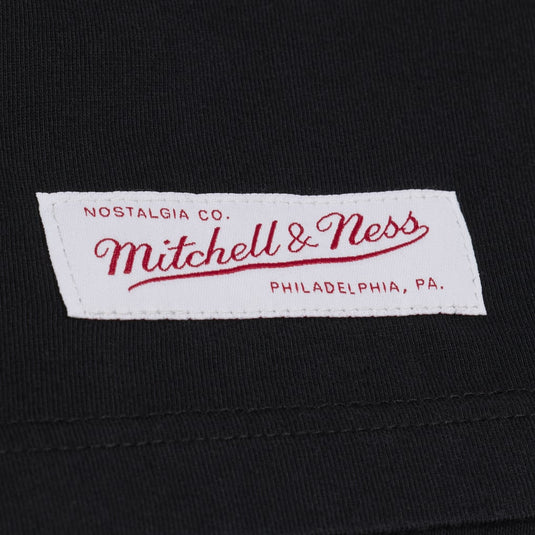 Close-up of a black knit garment with a white Mitchell & Ness brand label sewn onto the chest. The label features red embroidered text reading 'Nostalgia Co.
