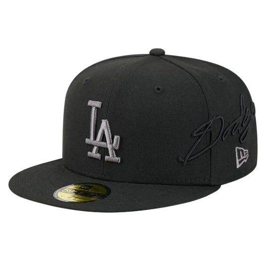 A black Los Angeles Dodgers fitted cap featuring a silver embroidered 'LA' logo on the front, a black autograph on the side, and a gold sticker on the brim.