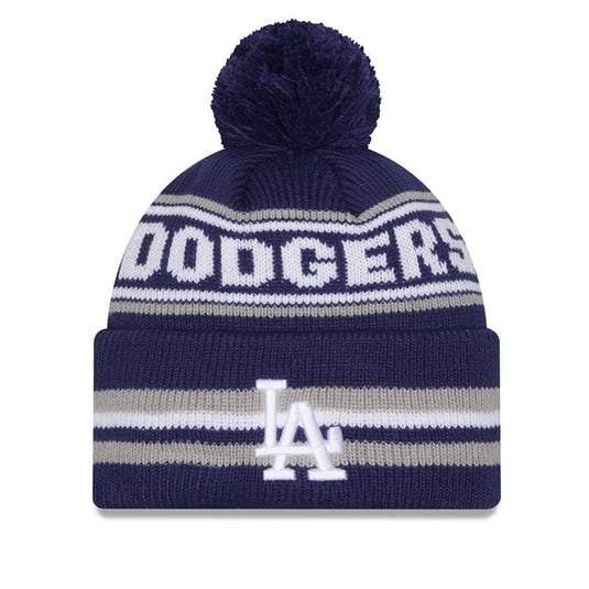 A navy blue knit pom-pom beanie with white and gray stripes, featuring the word 'DODGERS' across the top and the 'LA' Dodgers logo on the front cuff.