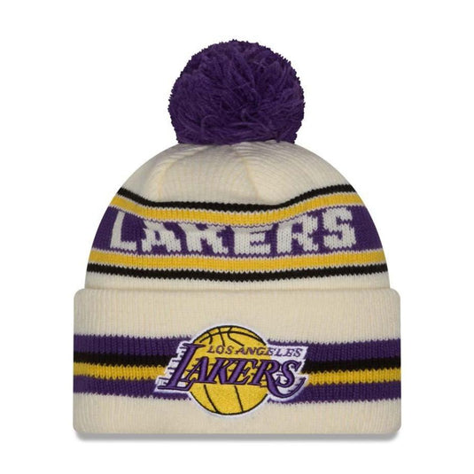 A knit pom-pom beanie hat featuring the Los Angeles Lakers logo and 'LAKERS' text in purple and yellow on a cream background, with a matching purple pom-pom on top.
