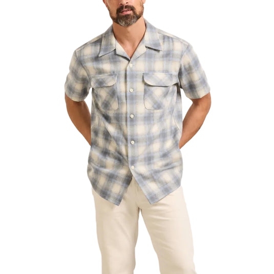 A man modeling a short-sleeve button-up plaid shirt in light blue and white, paired with off-white trousers and beige shoes, against a plain light beige studio background.