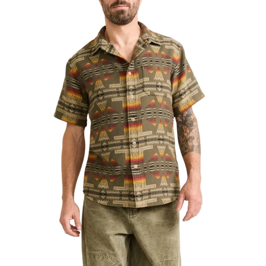 A man modeling a short-sleeve button-up shirt with a geometric Southwestern pattern in olive green, brown, and orange, paired with olive cargo pants and beige shoes, against a plain off-white studio background.