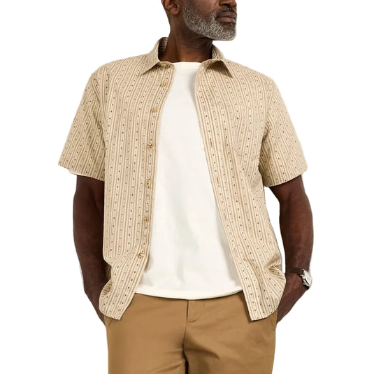 A man wearing a beige short-sleeve patterned button-up shirt over a white t-shirt, paired with tan trousers and off-white sneakers, standing against a plain off-white wall.