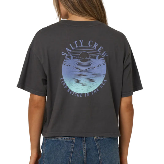 A woman viewed from the back wearing a dark charcoal gray cropped t-shirt with a large circular graphic on the back that reads 'SALTY CREW' and 'FIND REFUGE IN THE SEA'. She is wearing blue jeans against a solid light gray studio background.