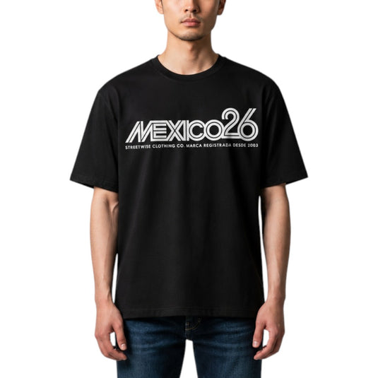 A man wearing a black short-sleeve t-shirt with a white 'MEXICO26' graphic on the chest, paired with blue jeans, against a plain white background.