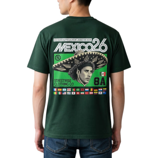 A man wearing a dark green t-shirt, viewed from the back. The shirt features a large graphic print with 'MEXICO26' in white, a green background, a large sombrero, a stylized portrait, and a row of small national flags at the bottom.