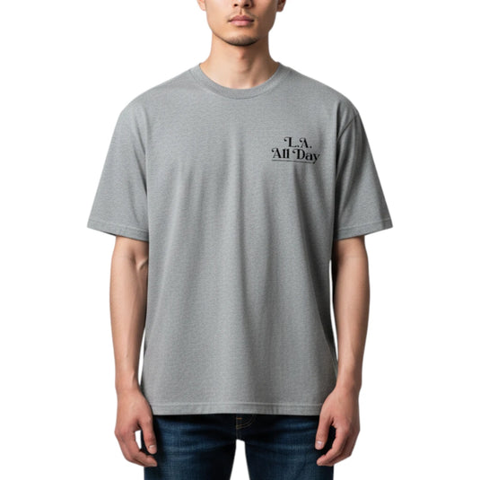 A man wearing a light gray crewneck t-shirt with a small black graphic on the left chest that reads 'L.A. All Day'.