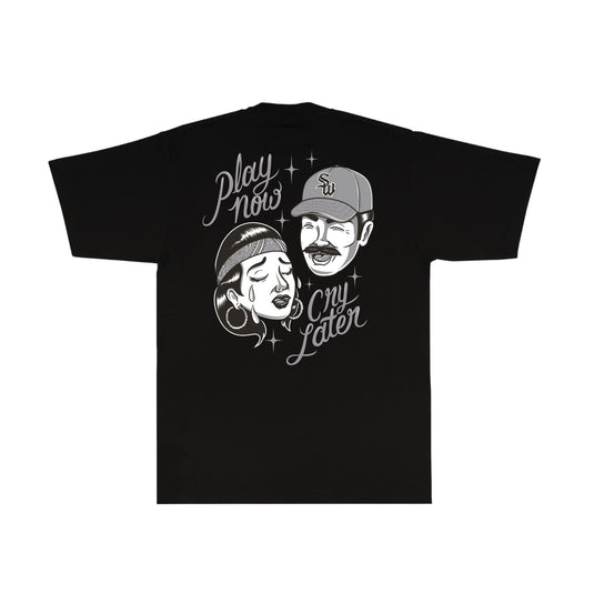 A black short-sleeve t-shirt with a white graphic print on the front, featuring a retro-style illustration of a man and woman wearing caps and bandanas, with the text 'Play now Cry later' and small star accents.