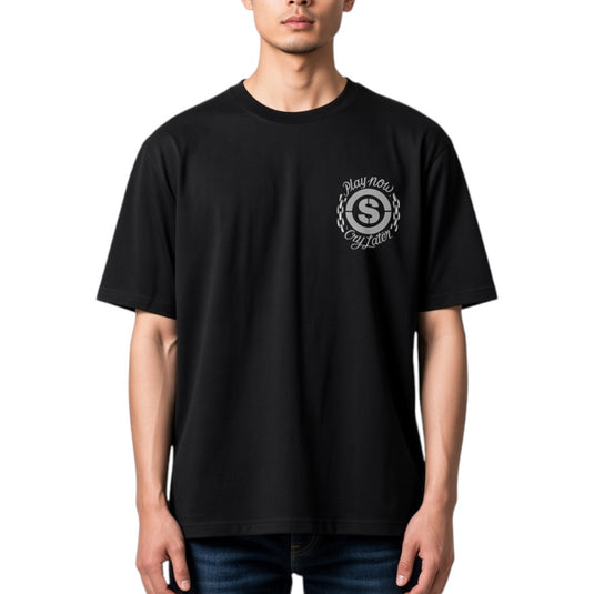 A man wearing a black short-sleeve crewneck t-shirt with a white circular graphic on the left chest featuring a stylized letter 'S' and the words 'Play Now Cry Later'