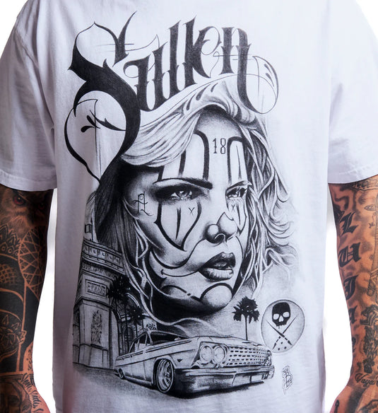 A close-up of a white short-sleeve t-shirt featuring a large black and gray graphic print of a stylized woman's face, the word 'Sullen' in gothic lettering, a classic car, and other street-art elements. The shirt is shown on a model with tattooed arms.