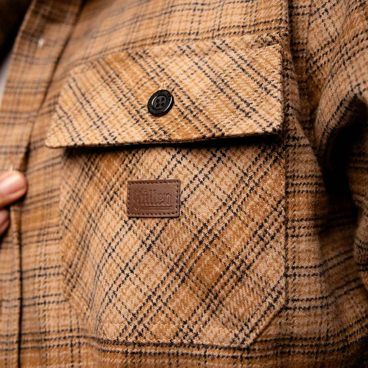 Close-up of a brown and tan plaid flannel shirt jacket pocket with a brown leather brand patch reading 'Sullen' and a black button.