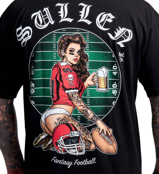 A close-up of a black graphic t-shirt featuring a large 'Fantasy Football' design by Sullen Art Co. The graphic shows a pin-up style illustration of a tattooed woman kneeling on a green football field background, holding a football and a beer mug, with a red helmet in front of her.
