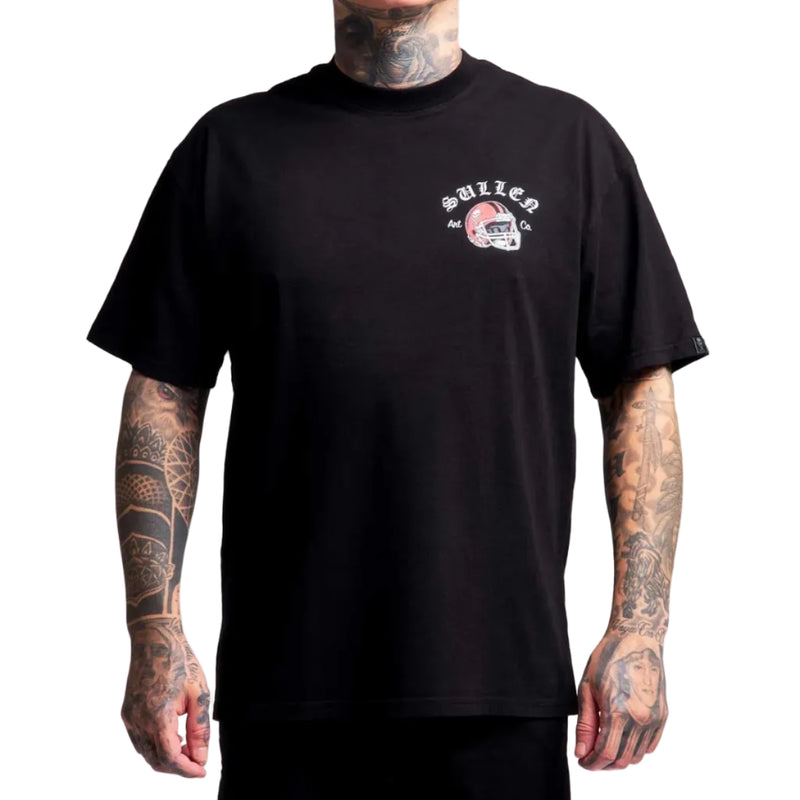 Load image into Gallery viewer, A man with visible tattoos wears a black short-sleeve t-shirt featuring a small white and red graphic on the left chest with the text 'sullen' and a football helmet illustration, set against a plain white background.
