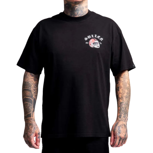 A man with visible tattoos wears a black short-sleeve t-shirt featuring a small white and red graphic on the left chest with the text 'sullen' and a football helmet illustration, set against a plain white background.