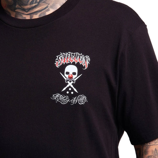 A close-up of a person wearing a black short-sleeve T-shirt. The shirt features a graphic on the chest with a white skull and crossed bones, a red clown nose, and stylized white and red text above and below.